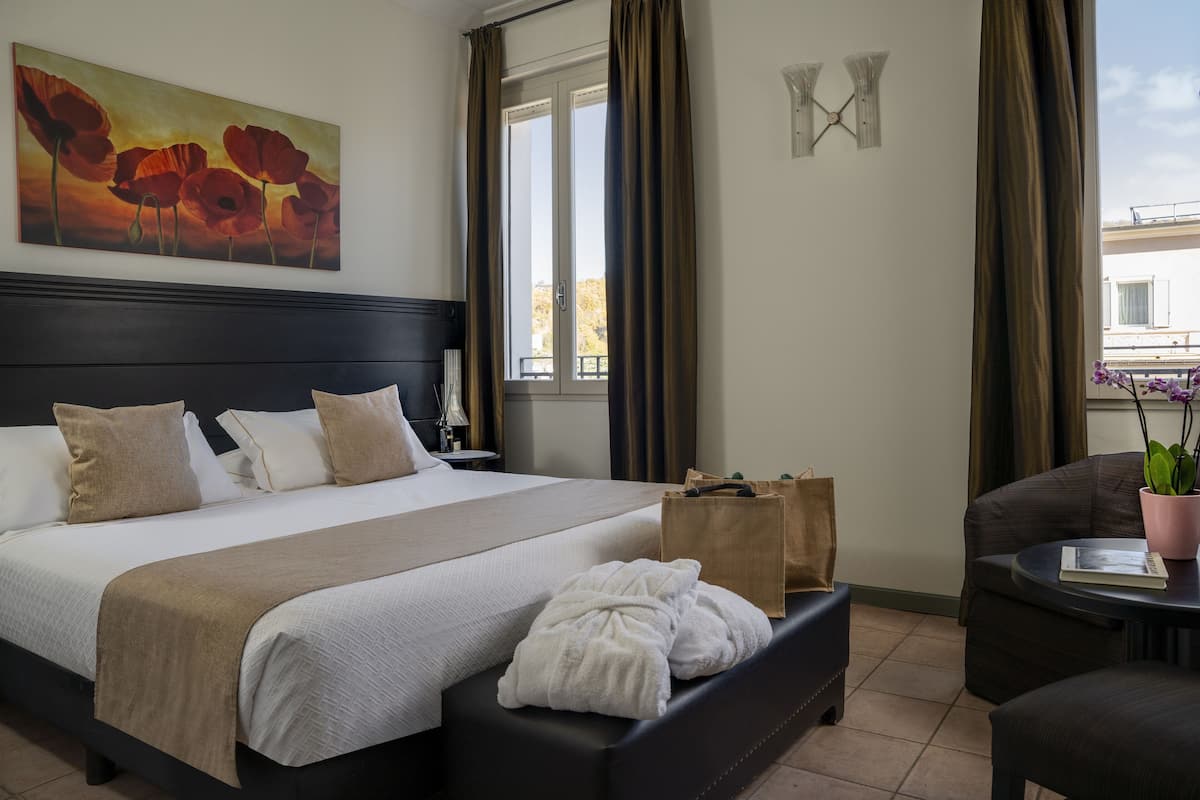 A hotel room with a bed, a chair, a table, a painting, and a window with curtains.