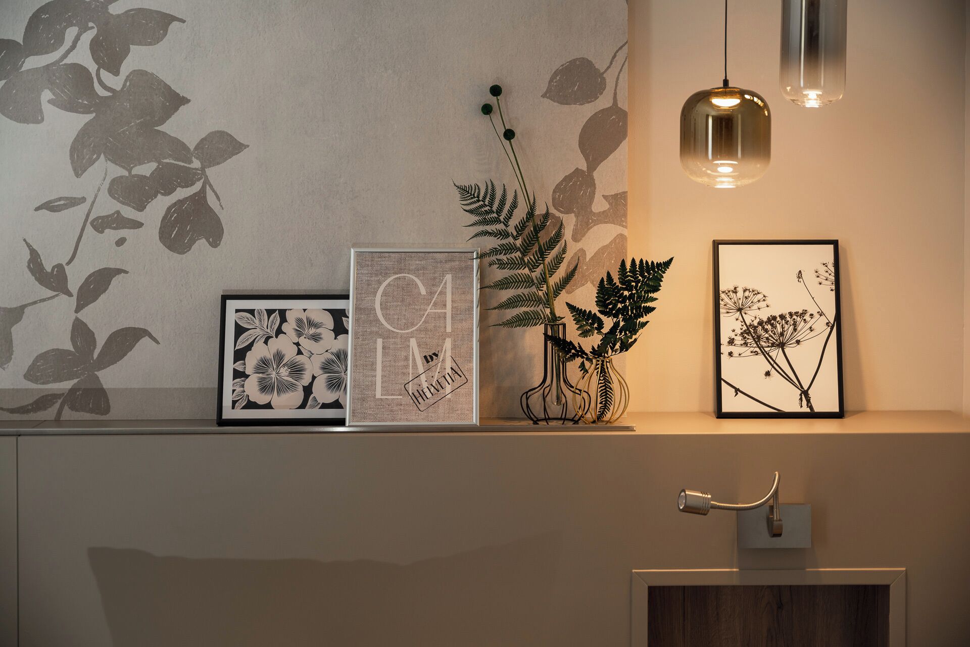 A bathroom with a wall-mounted shelf displaying framed botanical prints, a potted fern, and a wall-mounted light fixture.