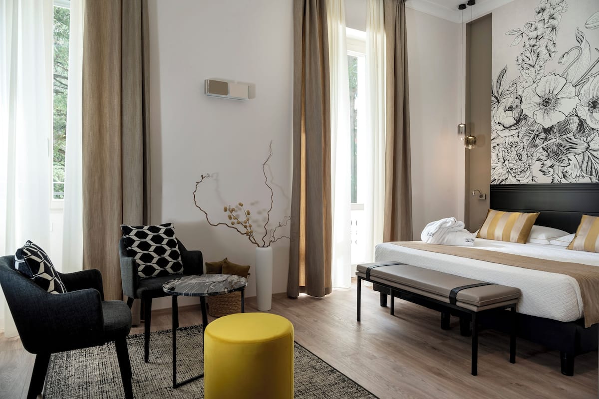 A modern hotel room with a large bed, a seating area with a bench, a chair, and a small table.