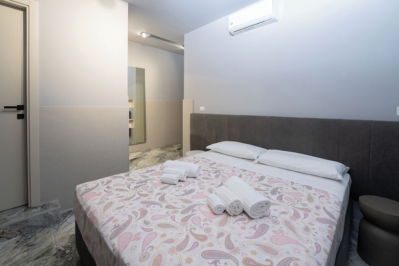 A hotel room with a bed, a headboard, a bedside table, and a bathroom visible in the background.