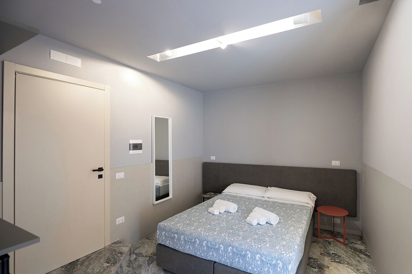 A bedroom with a bed, a bedside table, a mirror, and a door.