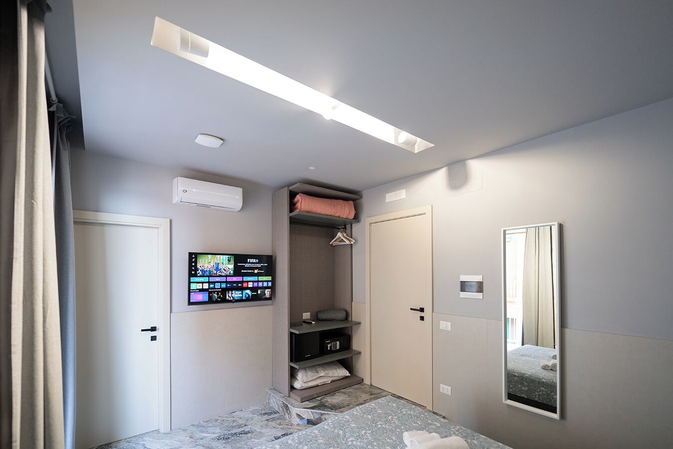 A hotel room with a bed, a TV mounted on the wall, a wardrobe, and a mirror.