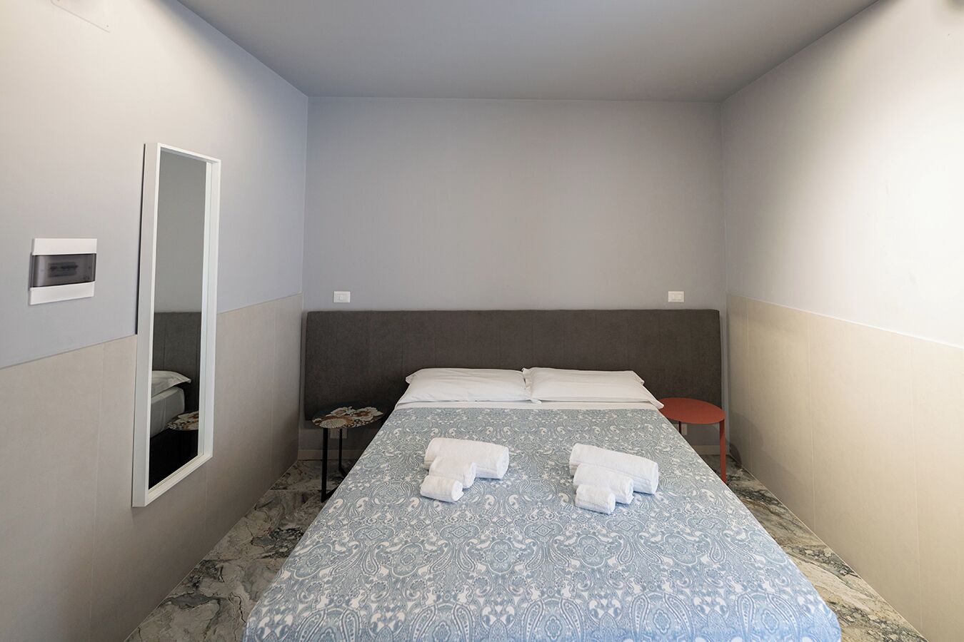 A small, modern hotel room with a single bed, a bedside table, a mirror, and a small red stool.