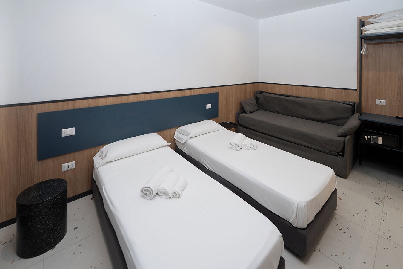 A hotel room with two single beds, a sofa, a small trash bin, and a wooden wardrobe.