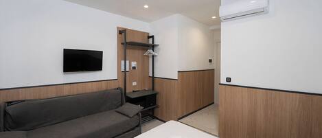 A modern hotel room with a grey sofa, a bed with white linens, a mounted TV, and an air conditioning unit.
