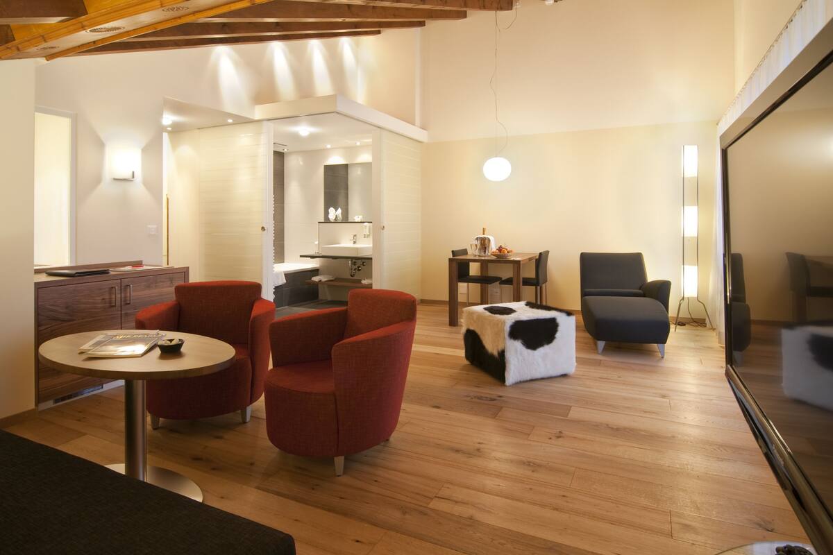 A modern living room with a wooden floor, red armchairs, a round table, a black and white ottoman, and a dining area with a small table and chairs.