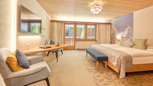 Un dormitorio acogedor con techo de madera, una cama grande con ropa de cama floral, un banco, un escritorio con silla y una ventana con cortinas.