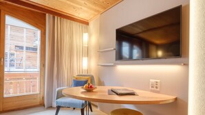 Una habitación con techo de madera, un televisor montado en la pared, una mesa redonda con una silla y una ventana con cortinas.
