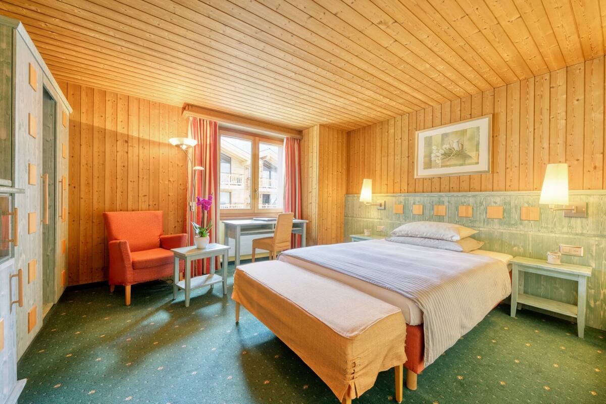 A wooden room with a bed, a chair, a table, and a window.