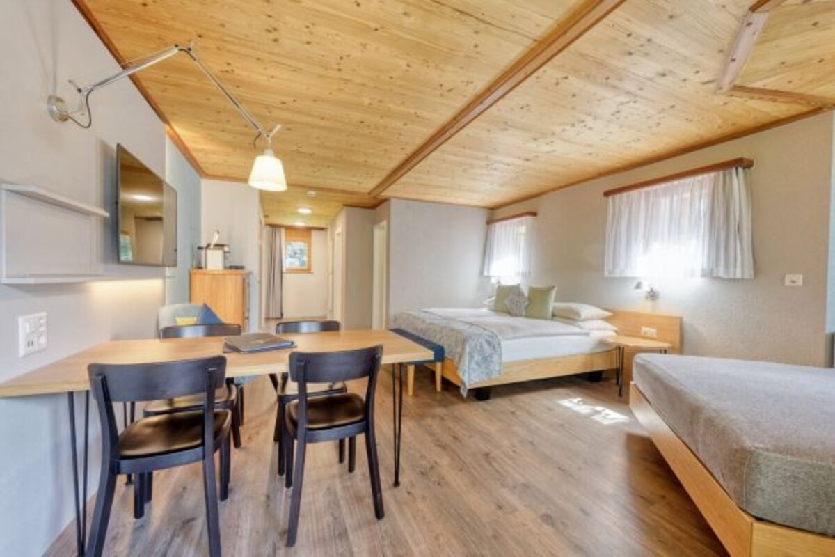 Een compacte kamer met een houten plafond, een bed, een bureau met stoelen en een raam met gordijnen.