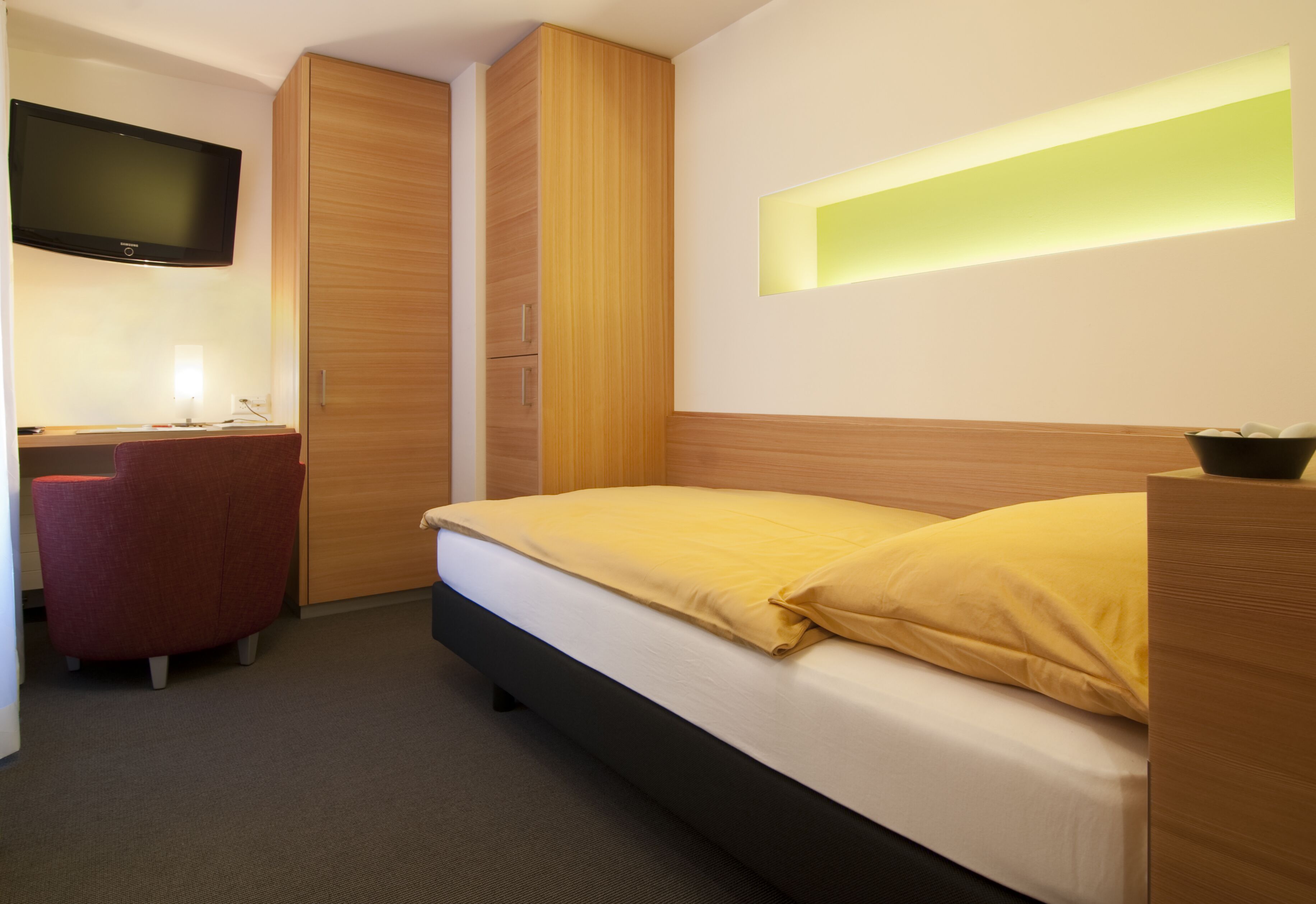 A hotel room with a bed, a desk, a chair, a TV, and a wall-mounted light.