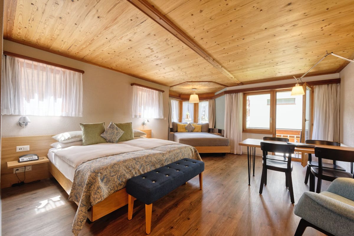A room with two beds, a desk, and a chair. There are windows with curtains, a bench, and a lamp.
