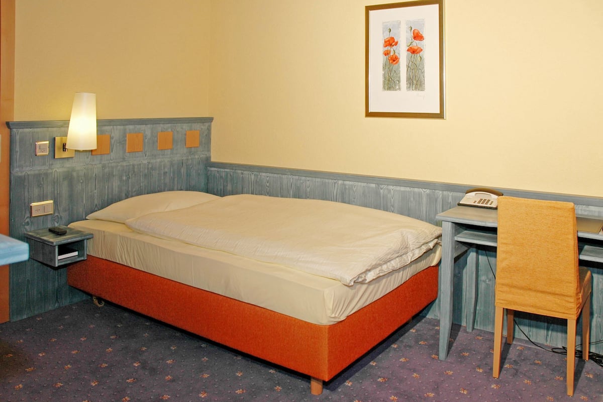 A hotel room with a bed, a desk, a chair, a lamp, and a framed picture on the wall.