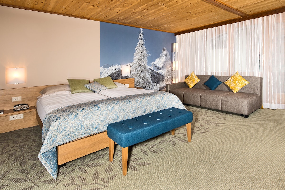 A bedroom with a bed, bench, sofa, and a painting of a snowy mountain.