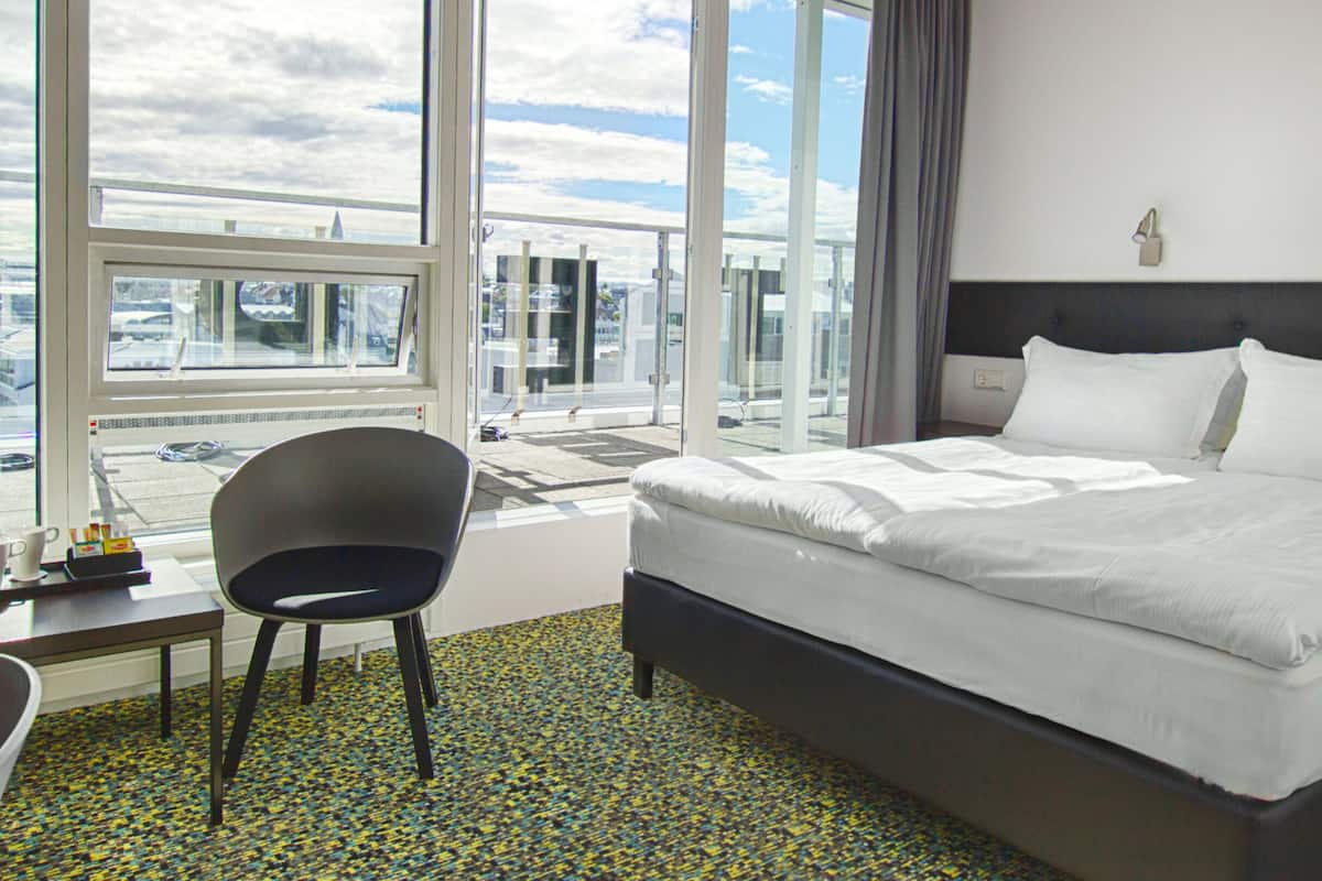 Een hotelkamer met een groot bed, een stoel, een bureau en uitzicht op de stad door grote ramen.