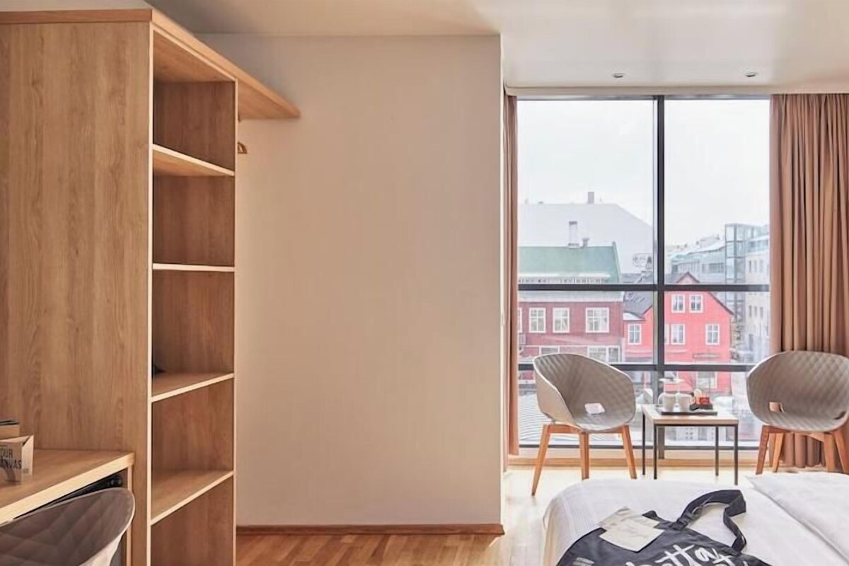A hotel room with a large window, a bed, a chair, a small table, and a wooden wardrobe.
