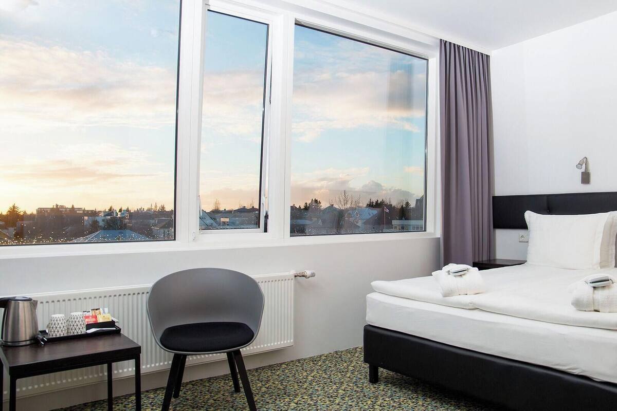 Een hotelkamer met een bed, een stoel, een tafel met waterkoker en een raam met uitzicht op de stad.