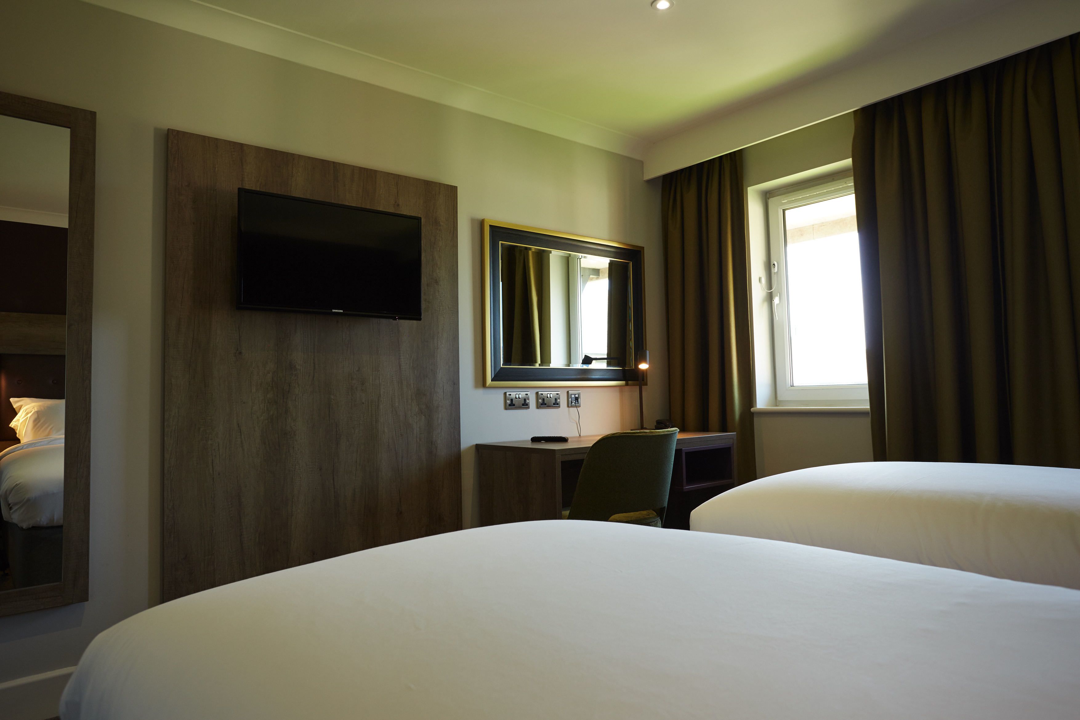 Ett hotellrum med två sängar, ett skrivbord, en stol, en platt-TV, en stor spegel och ett fönster med gardiner.
