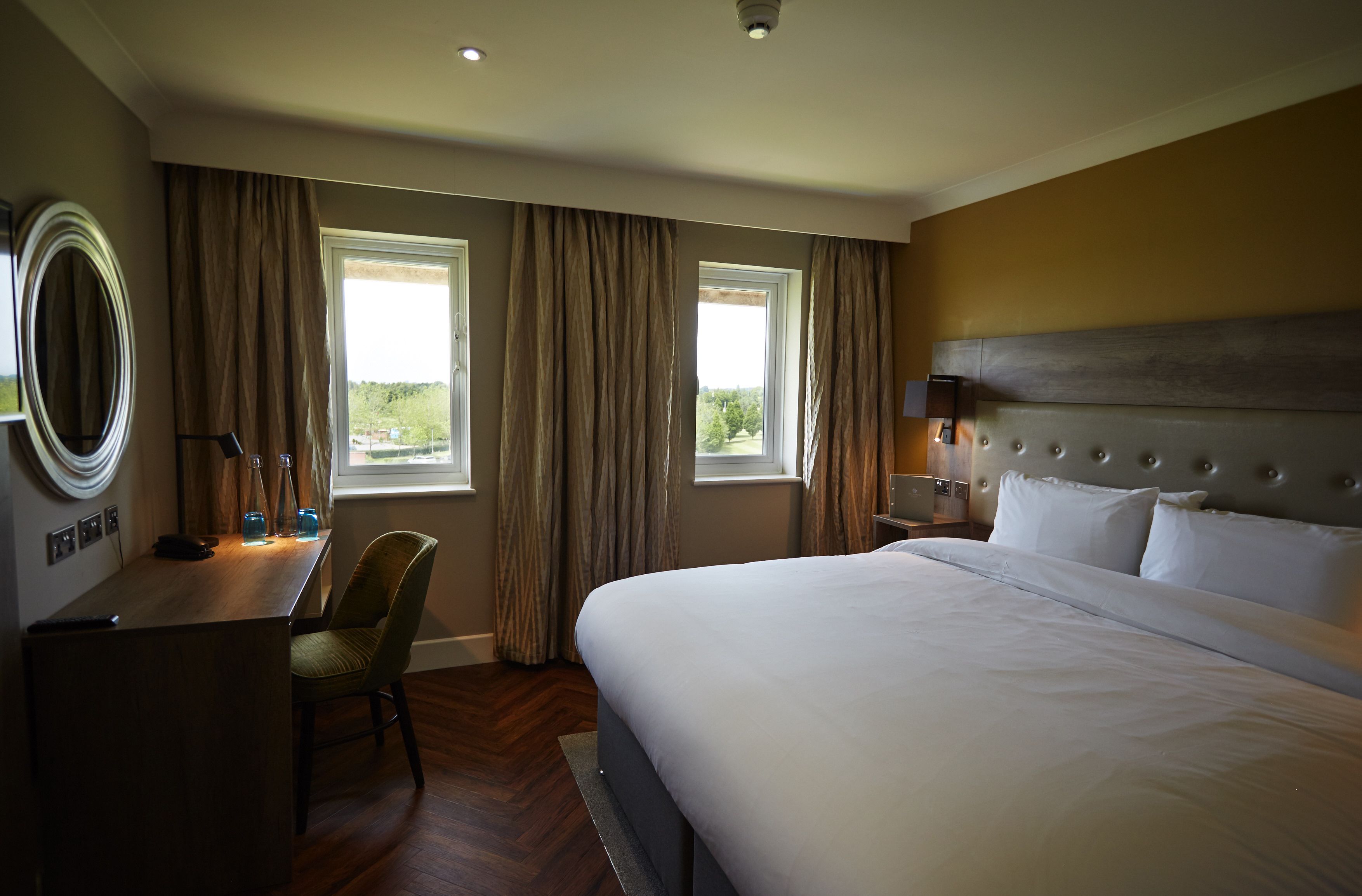 A hotel room with a bed, a desk, a chair, a nightstand, and a window with curtains.