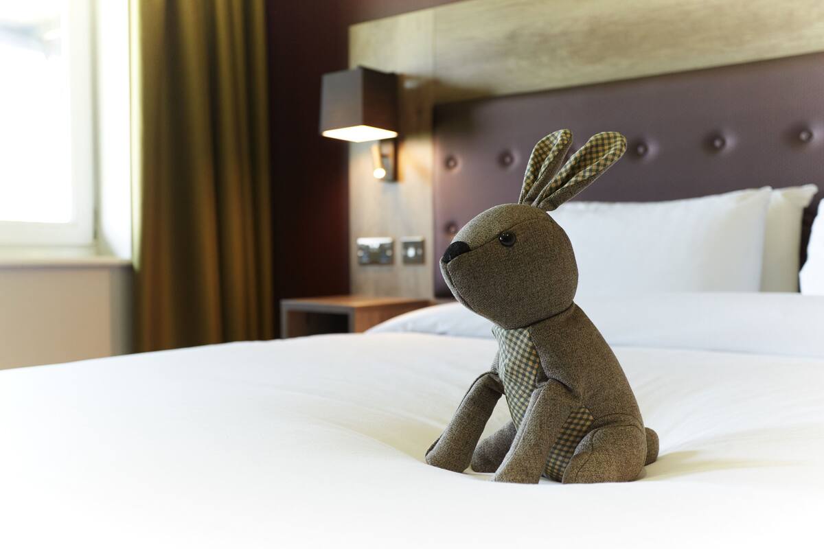 A plush toy on a bed in a hotel room with a tufted headboard and a bedside lamp.