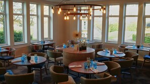 A restaurant with round tables, wicker chairs, and large windows offering views of the outdoors. - Wychwood Park (Crewe)