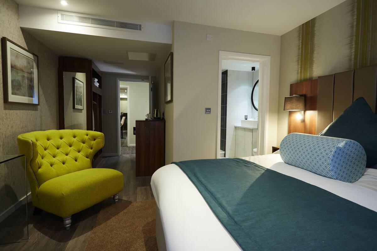 A hotel room with a bed, a yellow armchair, a mirror, and a door leading to a bathroom.