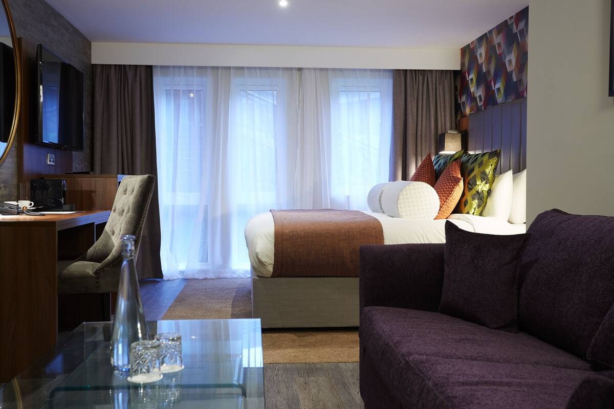 A modern hotel room with a bed, a sofa, a TV, and a desk with a coffee maker.