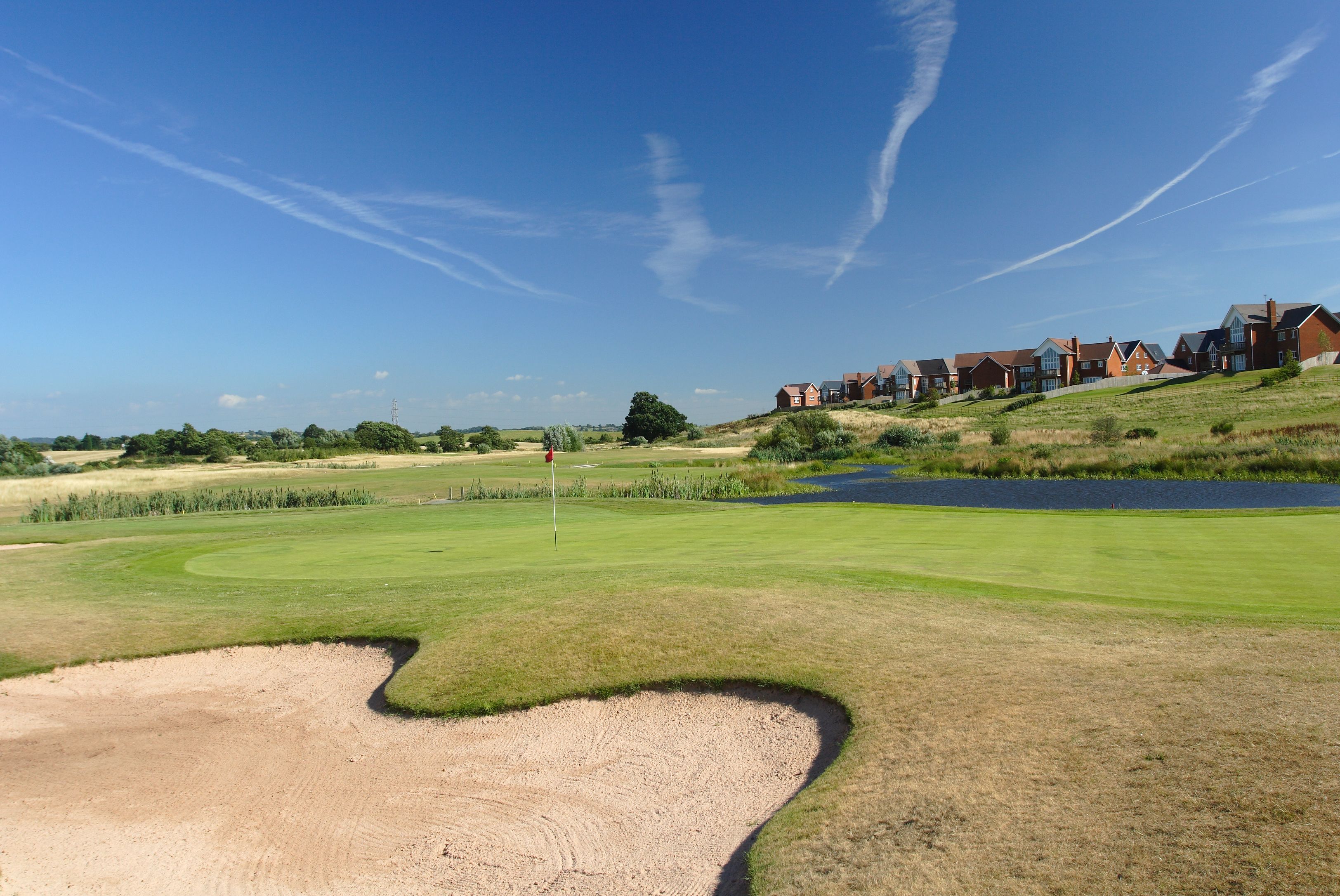 A golf course with bunkers, a pond, and a row of houses in the background.