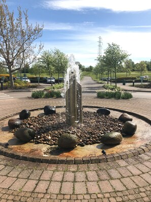 A fountain with water jets, surrounded by a circular arrangement of rocks and pebbles, set in a landscaped area with trees and a clear sky. - Wychwood Park (Crewe)