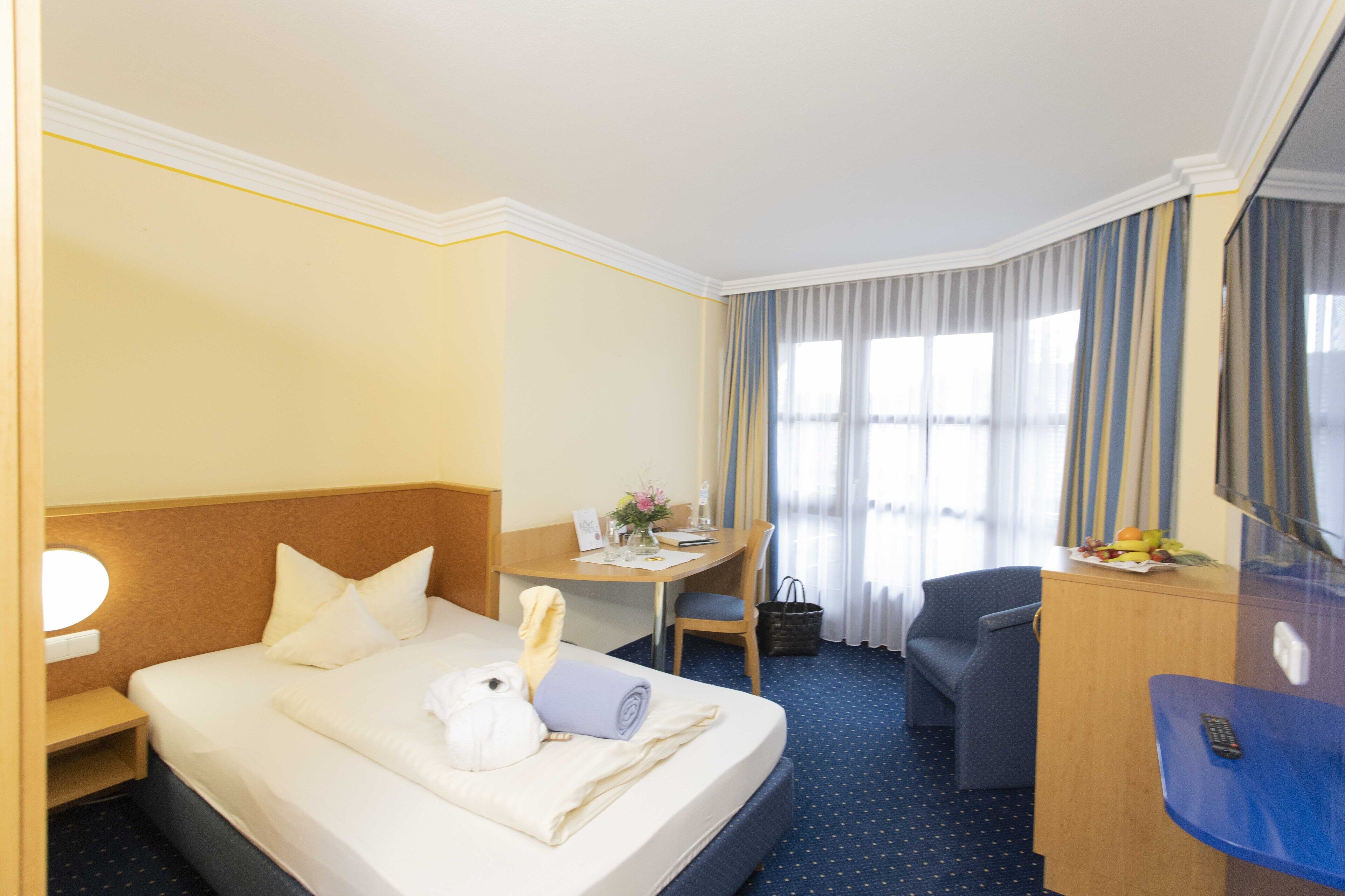A hotel room with a bed, a desk, a chair, a desk lamp, a small table with a fruit bowl, and a window with curtains.