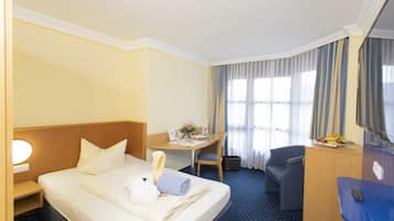 A hotel room with a bed, a desk, a chair, a desk lamp, a small table with a fruit bowl, and a window with curtains.