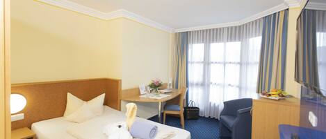 A hotel room with a bed, a desk, a chair, a desk lamp, a small table with a fruit bowl, and a window with curtains.