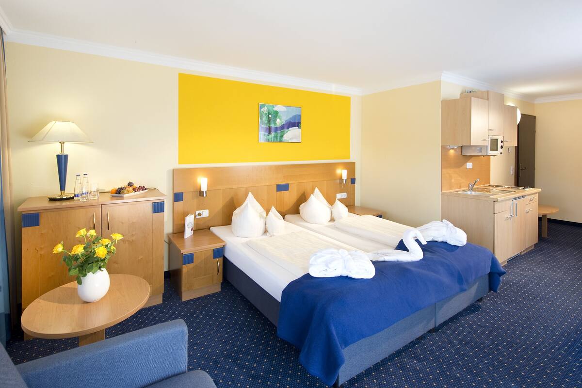 Une chambre d’hôtel avec un lit, un bureau, une chaise, une petite table sur laquelle est posé un vase de fleurs, une lampe et un tableau au mur.