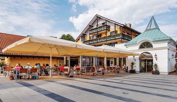 An outdoor seating area with tables and chairs under a large umbrella, adjacent to a building with balconies and a distinctive green roof. - Hotel Schmelmer Hof (Bad Aibling)