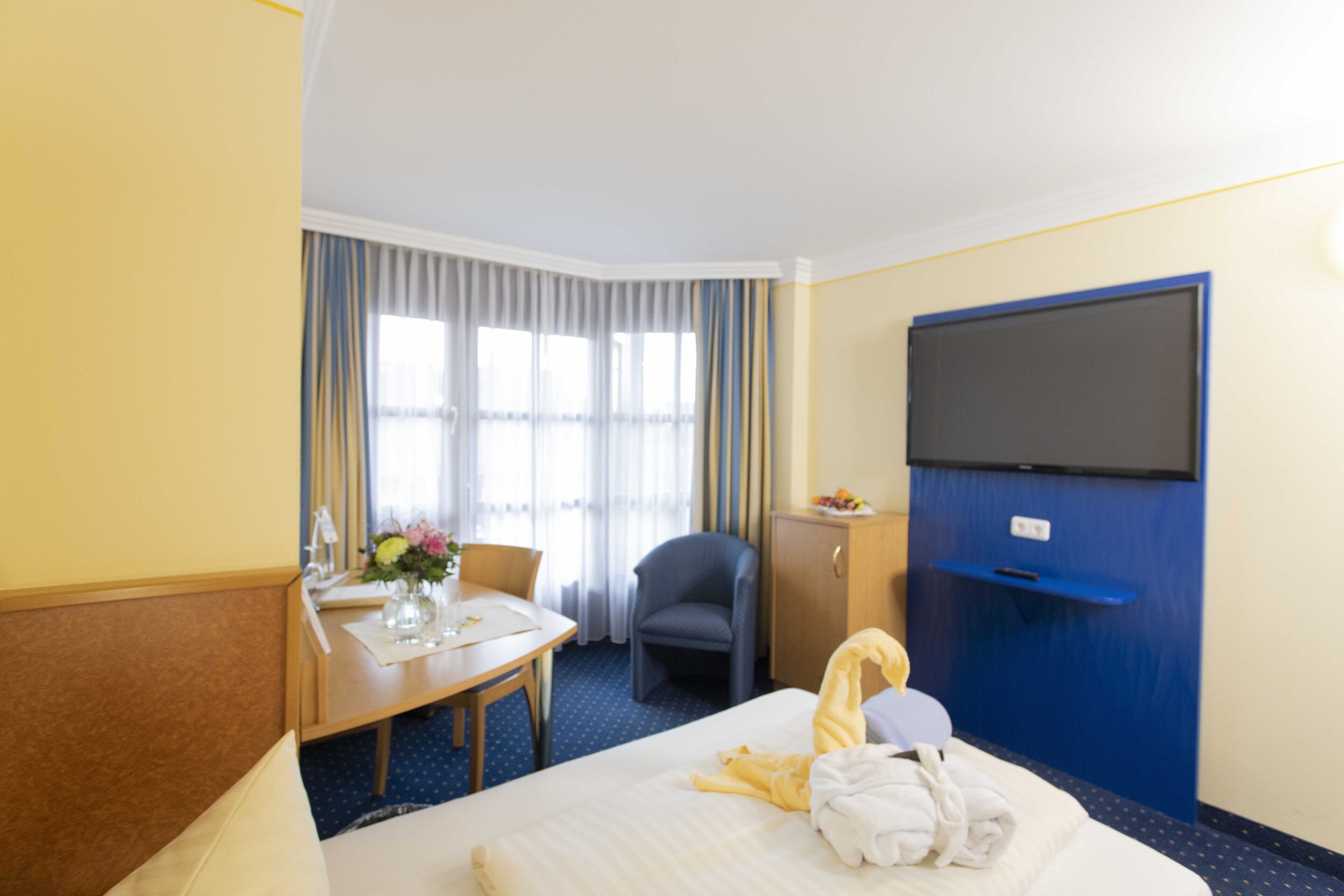 Une chambre d’hôtel comprenant un lit, un bureau avec une chaise, une télévision et une fenêtre avec des rideaux.