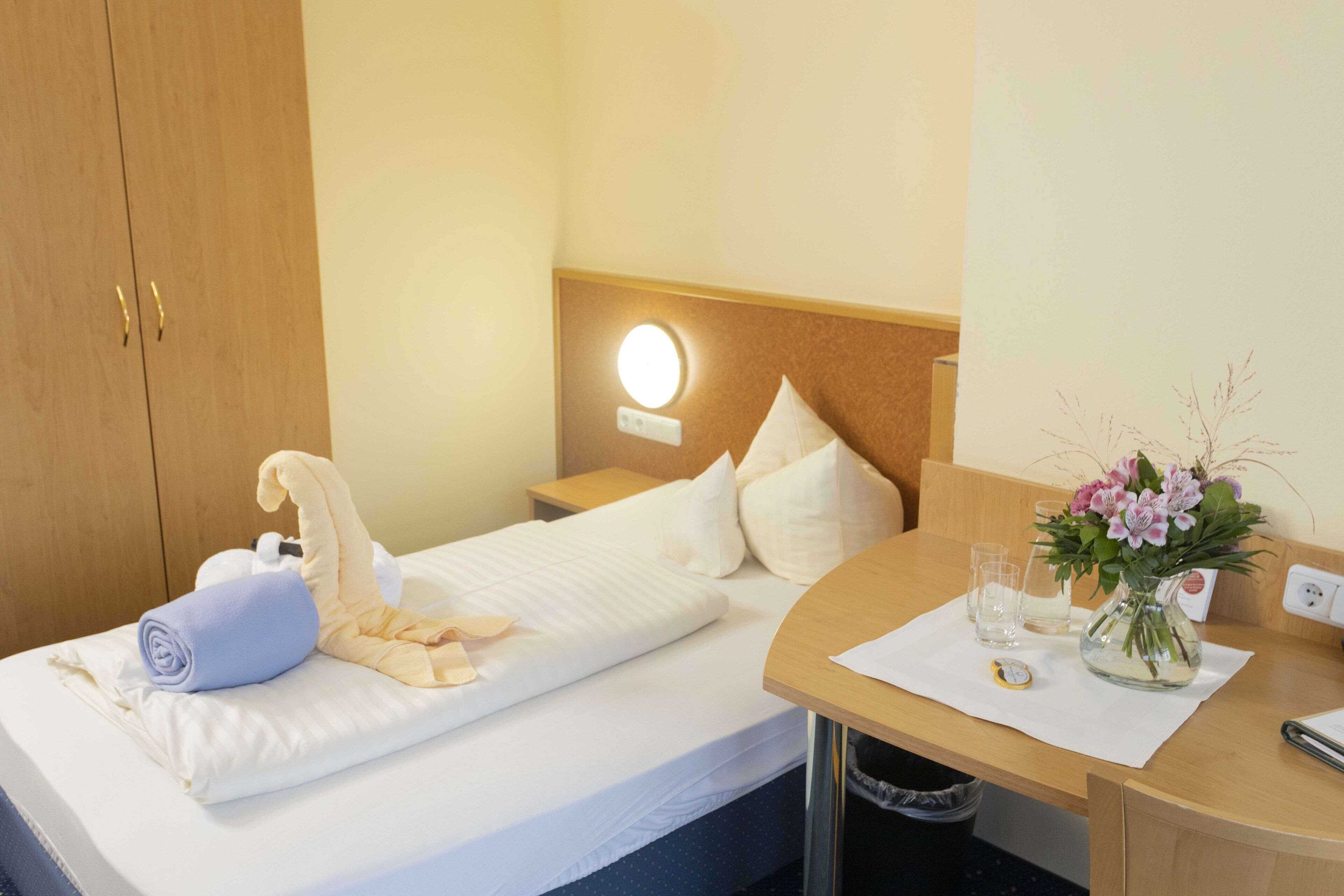 A neatly made bed with a rolled towel and a pillow, a bedside table with a vase of flowers, and a nightstand with a lamp.