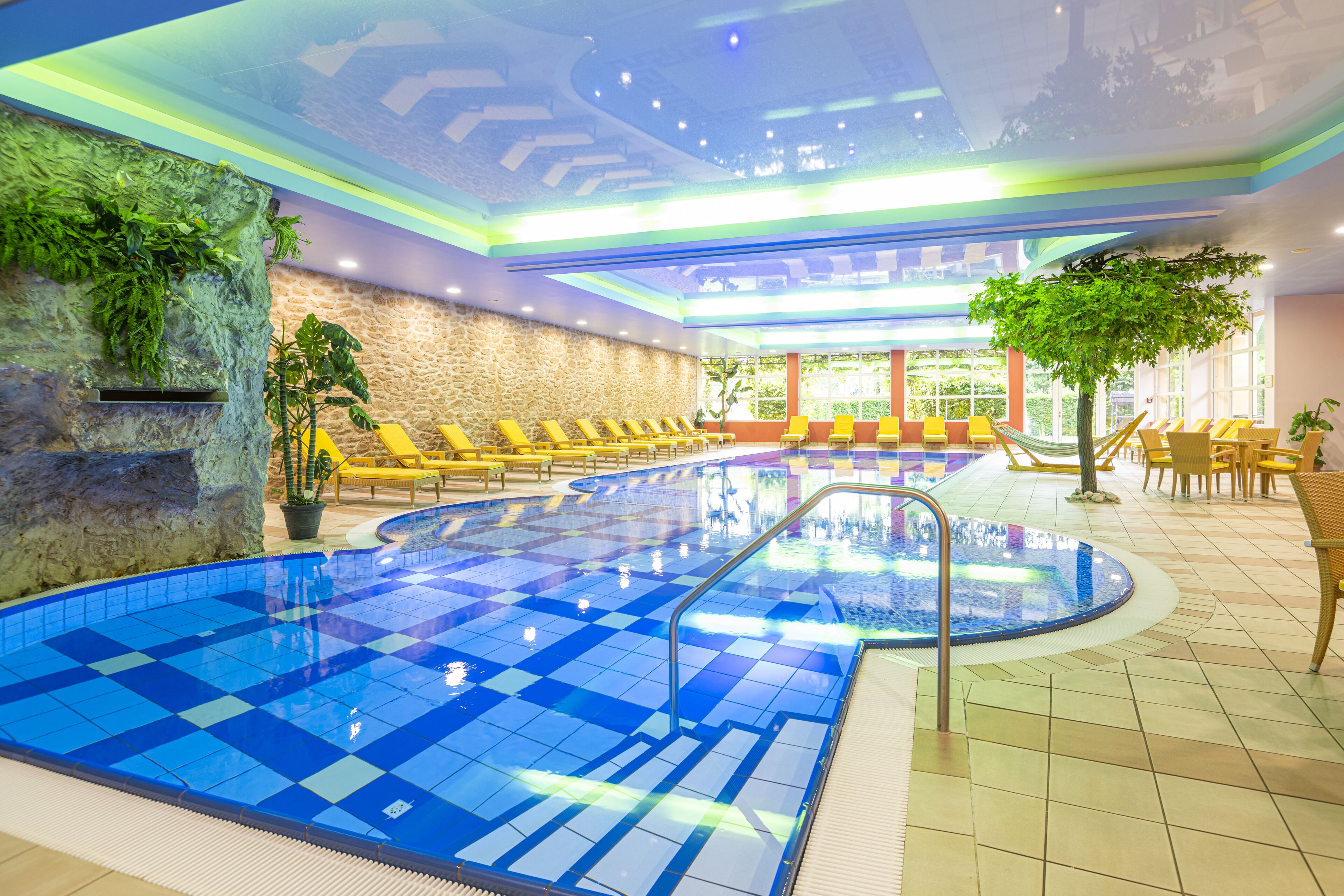An indoor swimming pool with blue tiles, yellow lounge chairs, and a stone wall with greenery.