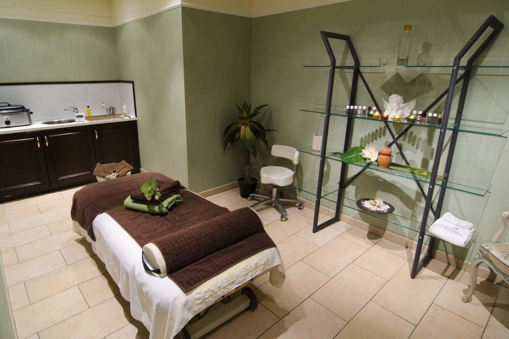 A spa room with a massage table, a small sink, a chair, a potted plant, and a glass shelf with toiletries.