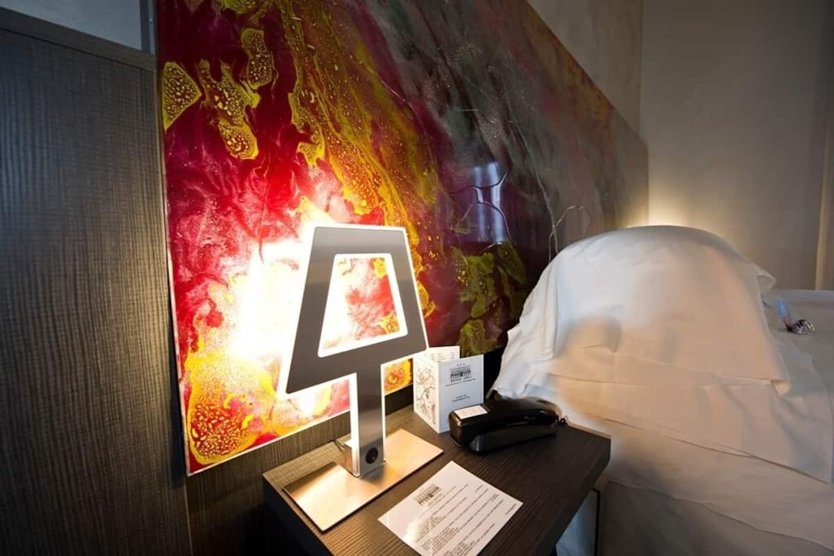 A hotel room with a bed, a bedside table with a lamp, a painting, and a wall-mounted phone.