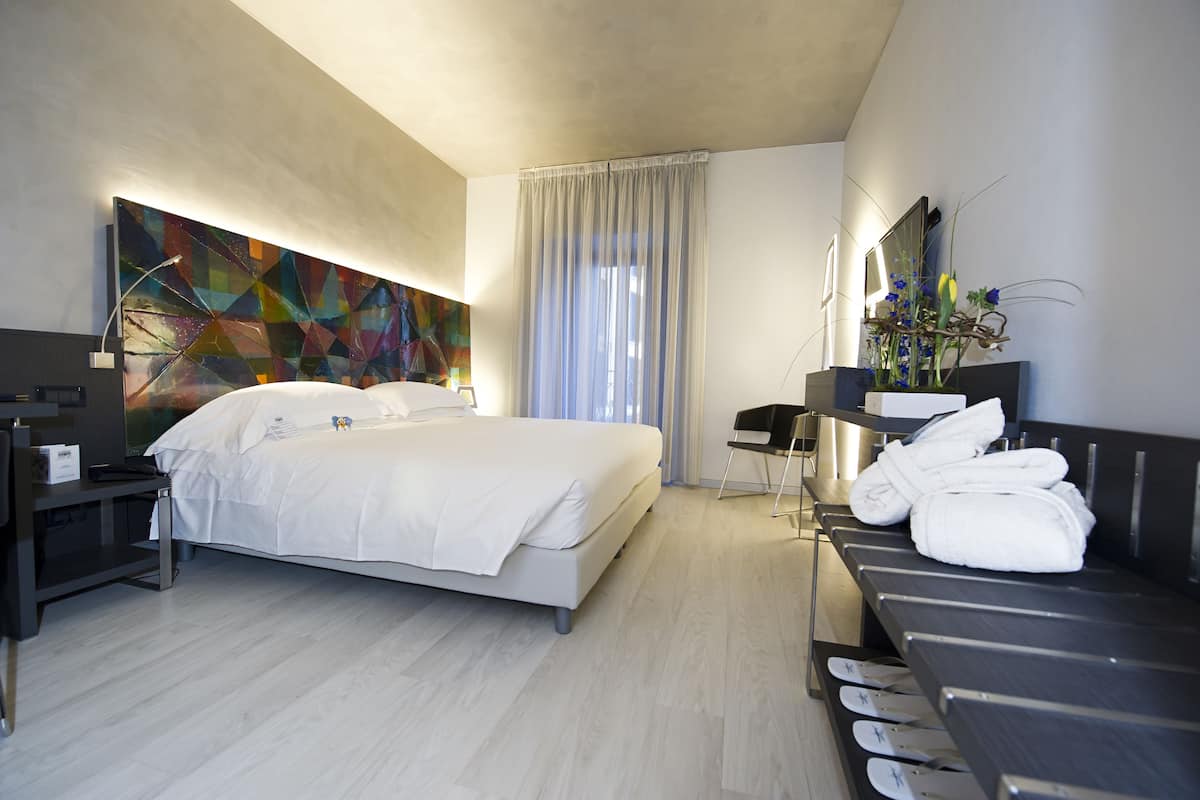A modern hotel room with a large bed, a desk with a chair, a vanity with a mirror, and a bathroom with a shower.