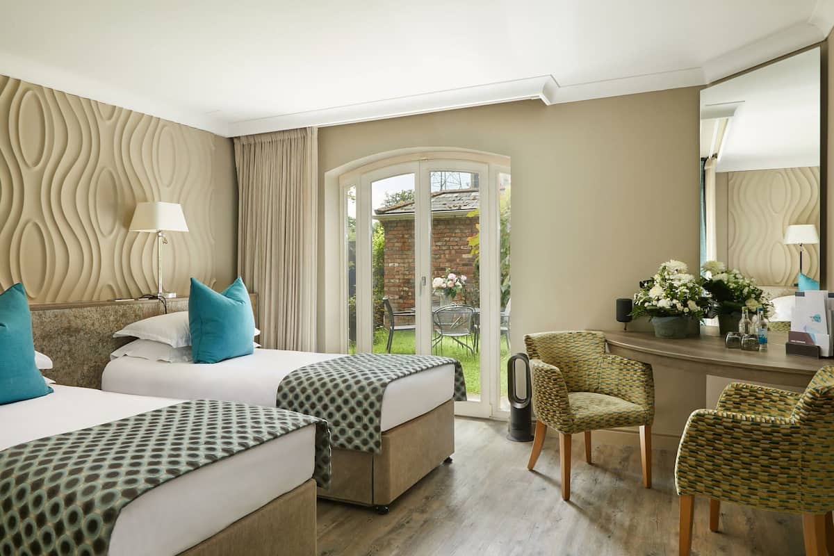 A hotel room with two beds, a sitting area with a chair, a desk with a lamp, and a view of the garden through a glass door.