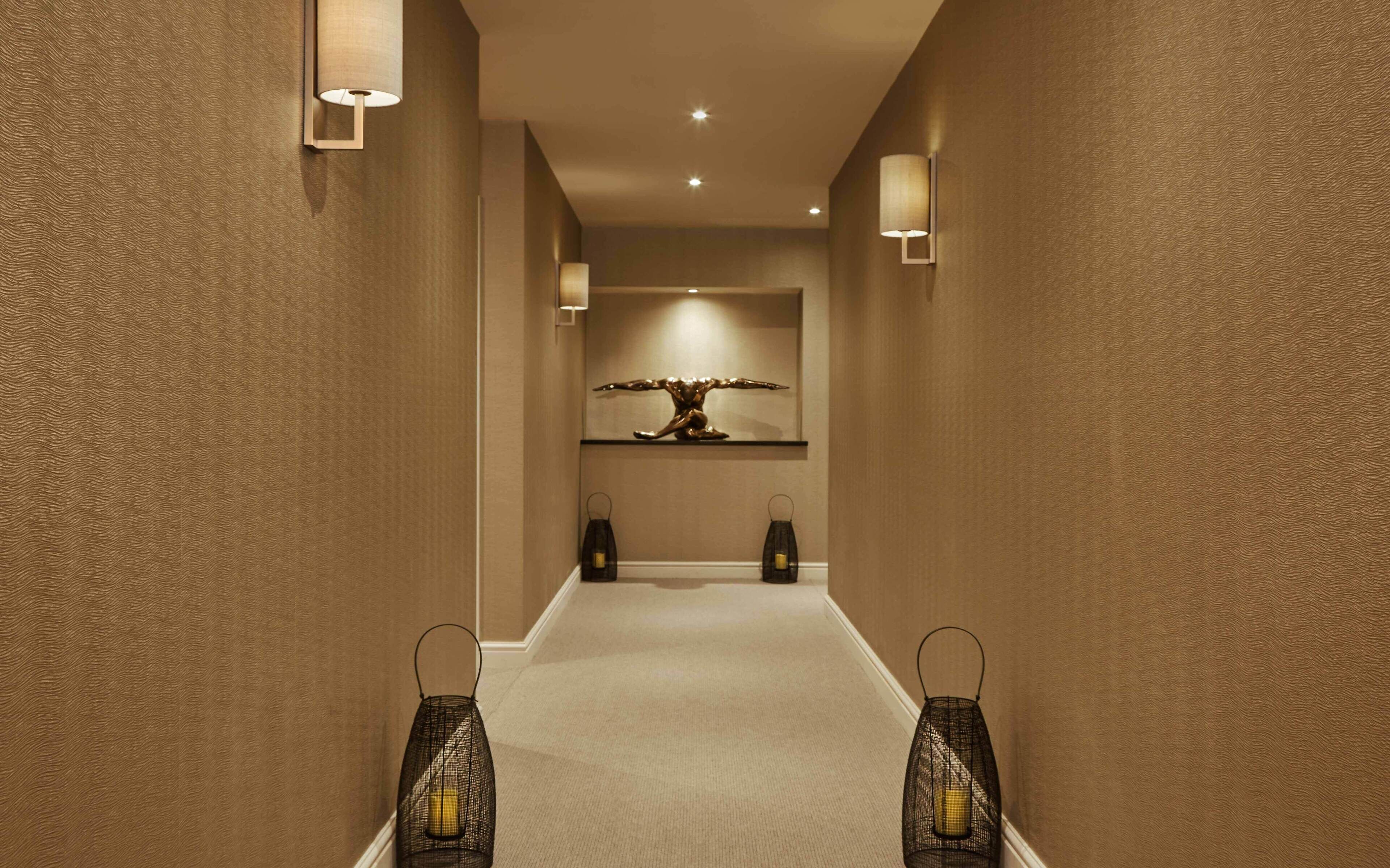 A hallway with textured walls, recessed ceiling lights, a central art piece, and two wall-mounted lanterns.