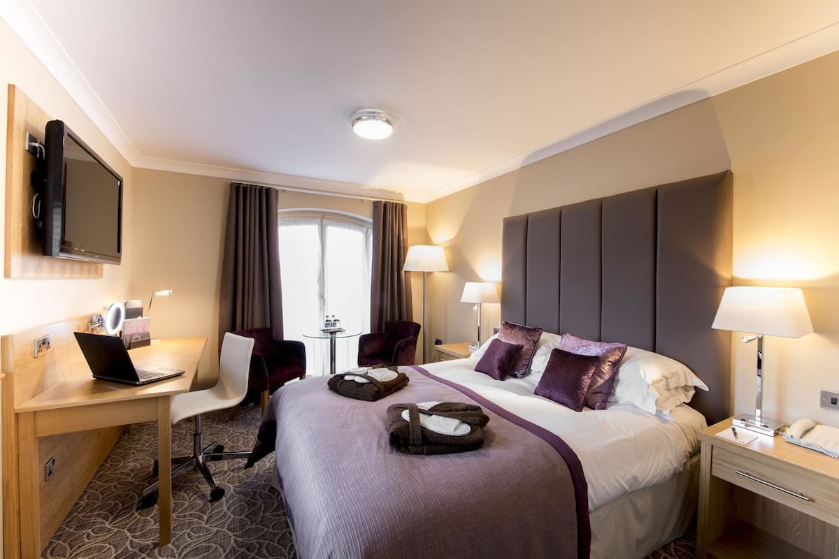 A hotel room with a bed, desk, chair, TV, lamps, and a window with curtains.