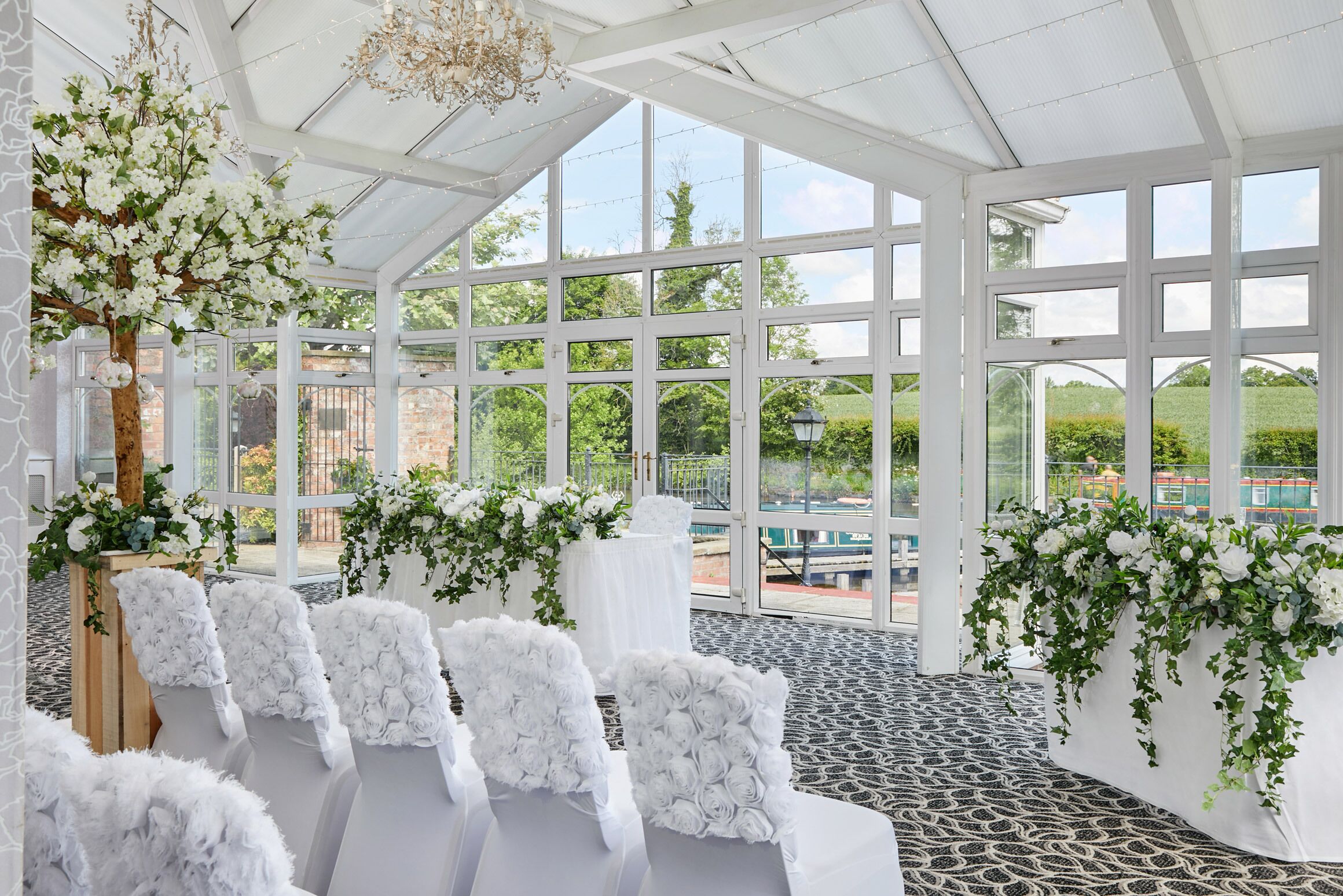 A conservatory with white floral arrangements, seating, and a view of greenery outside.
