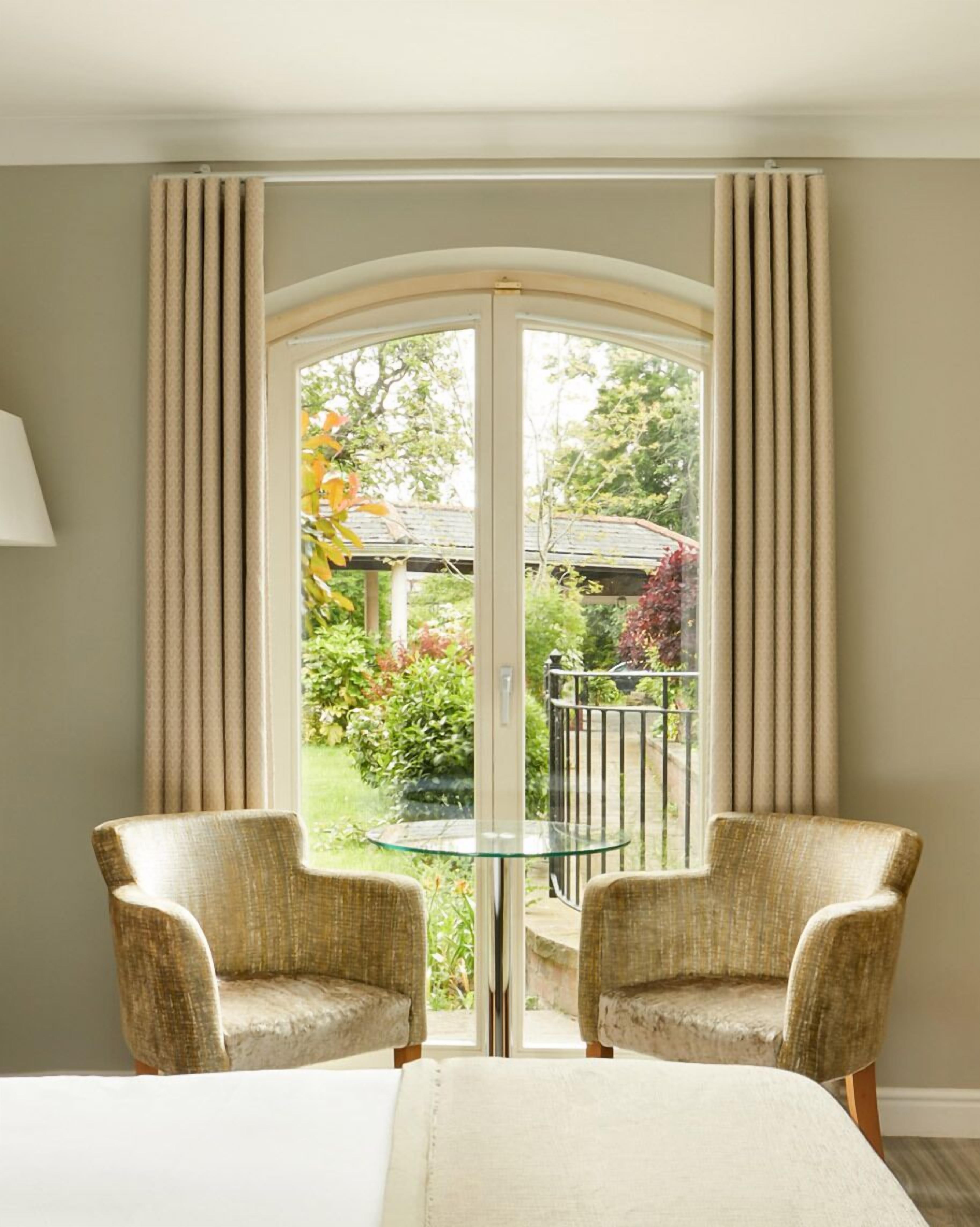 A room with a glass door leading to a garden, two wicker chairs, a small glass table, and a bed with a white headboard.