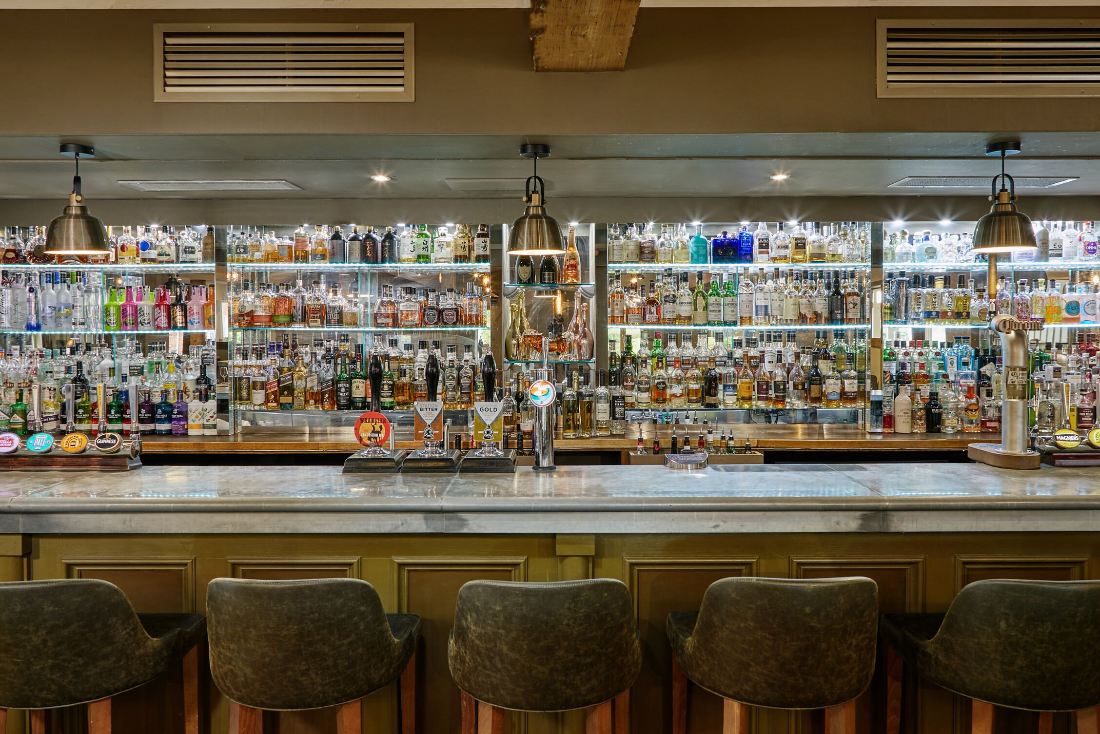 Un bar con varias estanterías llenas de botellas de licores, una barra con grifos y taburetes de bar.