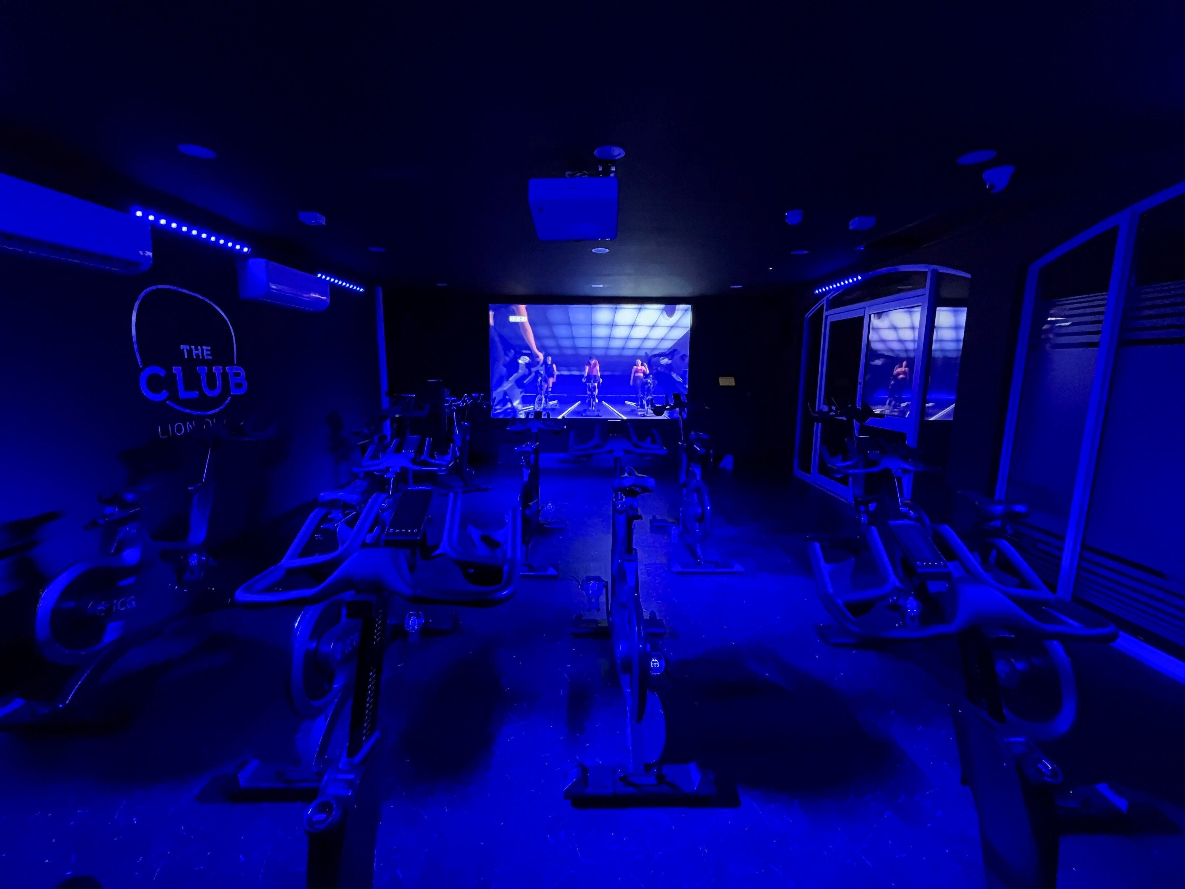 A gym with stationary bikes, a large screen displaying a workout video, and a neon sign reading "The Club".