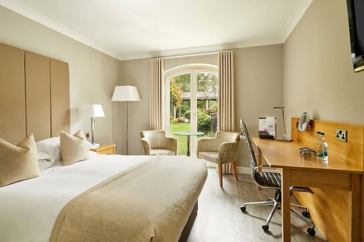 A hotel room with a large bed, a desk with a chair, a lamp, and a view of the outdoors.