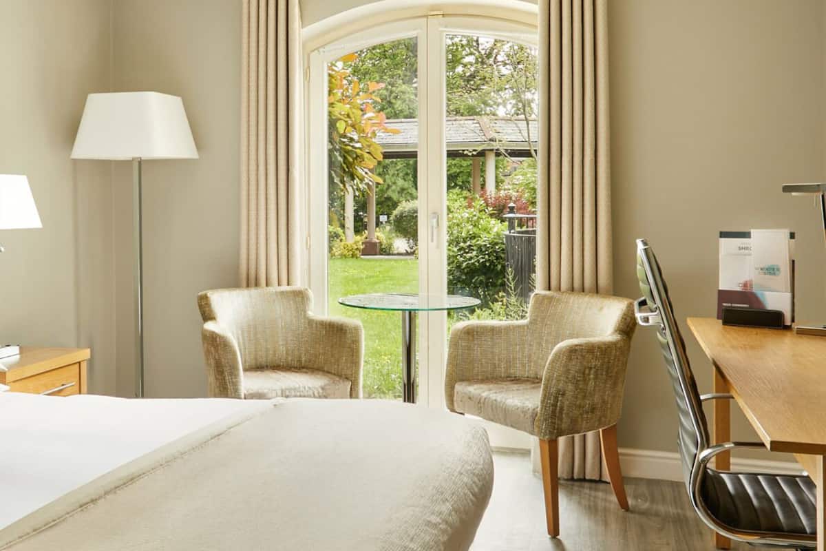 A hotel room with a bed, two armchairs, a desk with a chair, a lamp, and a view of a garden through a large window.