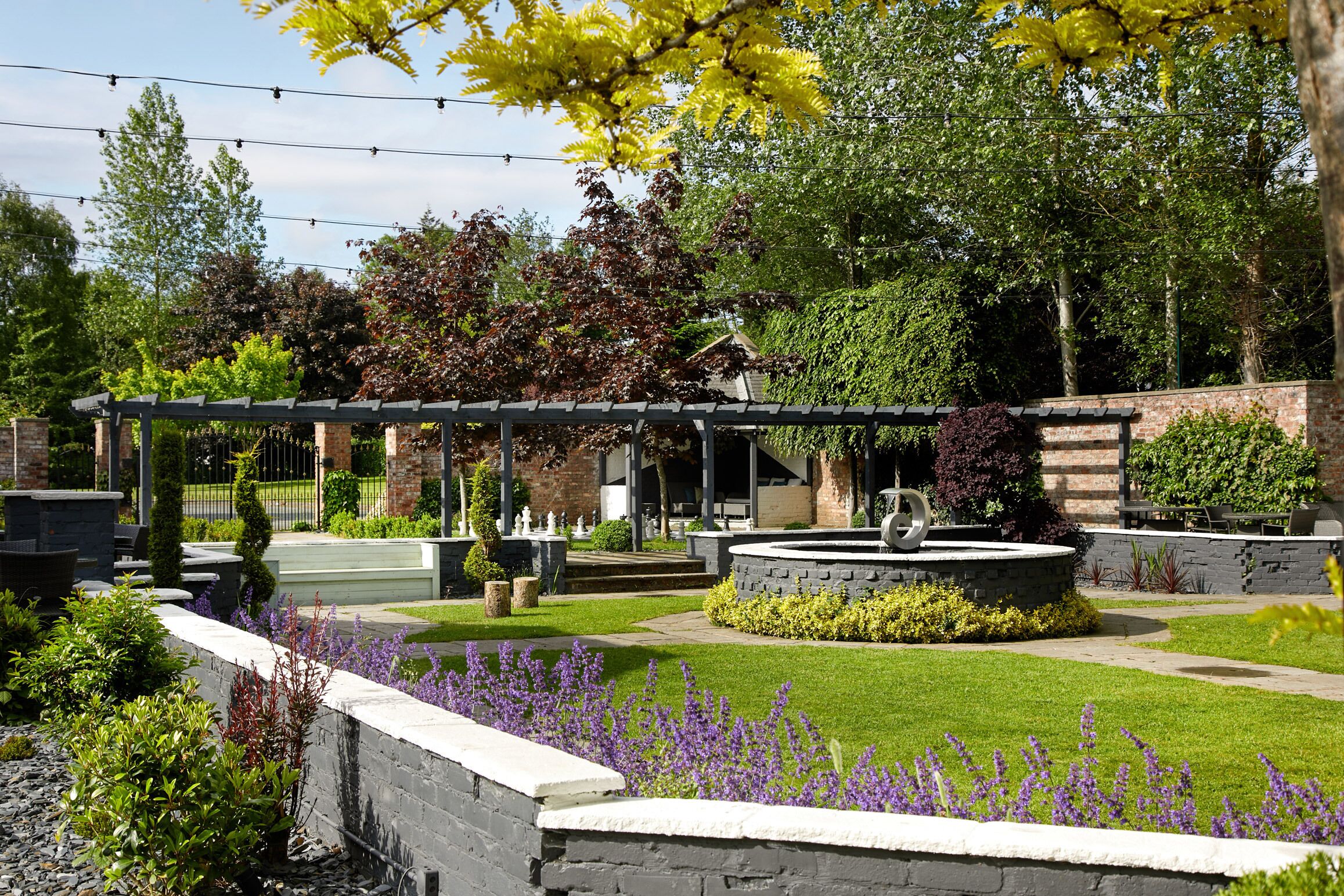 A landscaped garden with a pergola, a circular stone planter, and a variety of shrubs and flowers.
